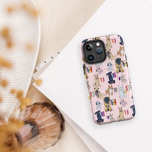Puede incluir: Una funda de teléfono rosa con un patrón repetido de varias razas de perros, cada una sosteniendo una bandera arcoíris. La funda tiene un módulo de cámara negro. El fondo es neutro con papel blanco.