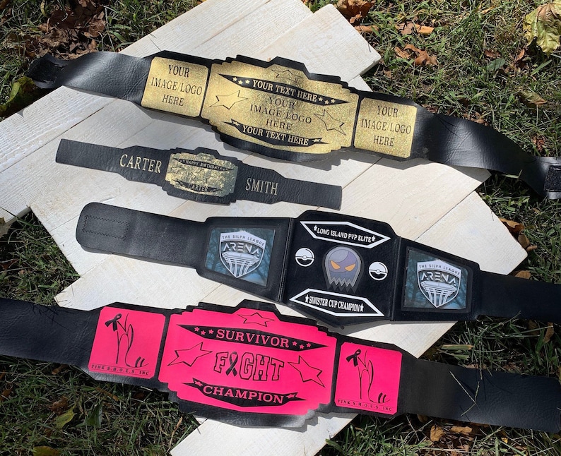 May include: Three black wrestling championship belts with gold and pink accents. The belts have different designs, including a ghost, a building, and a star. The belts are labelled "Long Island PVP Elite", "Sinister Cup Champion", "Survivor Fight Champion", and "The Silk League Arena".