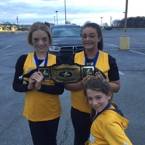 May include: Three young people wearing yellow shirts and black trousers are holding a gold championship belt with the words "Swann Mills Jr. High Playmaker" on it. They are standing in a car park.