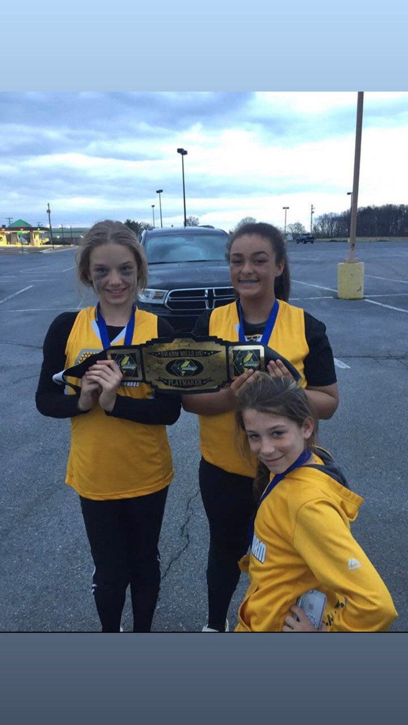 May include: Three young people wearing yellow shirts and black trousers are holding a gold championship belt with the words "Swann Mills Jr. High Playmaker" on it. They are standing in a car park.