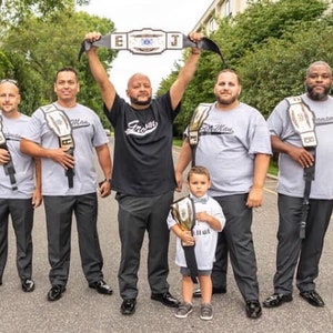 May include: A group of people, including children, are standing in a line holding championship belts. The belts are gold and black with white lettering. The people are wearing grey and black shirts with the words "Team Man" and "Forever" printed on them.