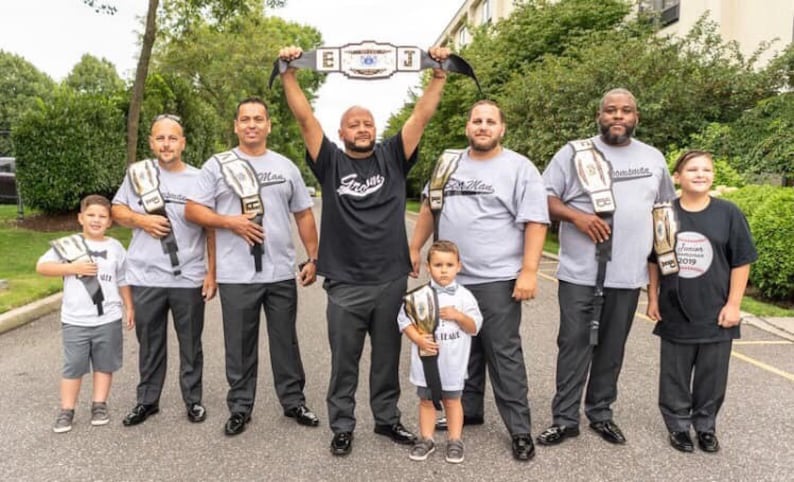 May include: A group of people, including children, are standing in a line holding championship belts. The belts are gold and black with white lettering. The people are wearing grey and black shirts with the words "Team Man" and "Forever" printed on them.