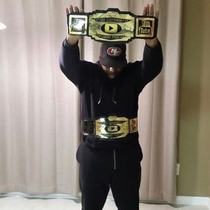 May include: A person wearing a black hoodie and black trousers is holding a gold championship belt with the words "100K Subscribers" and "YouTube" on it. The belt also has a play button icon and the words "Lamont Tyson".