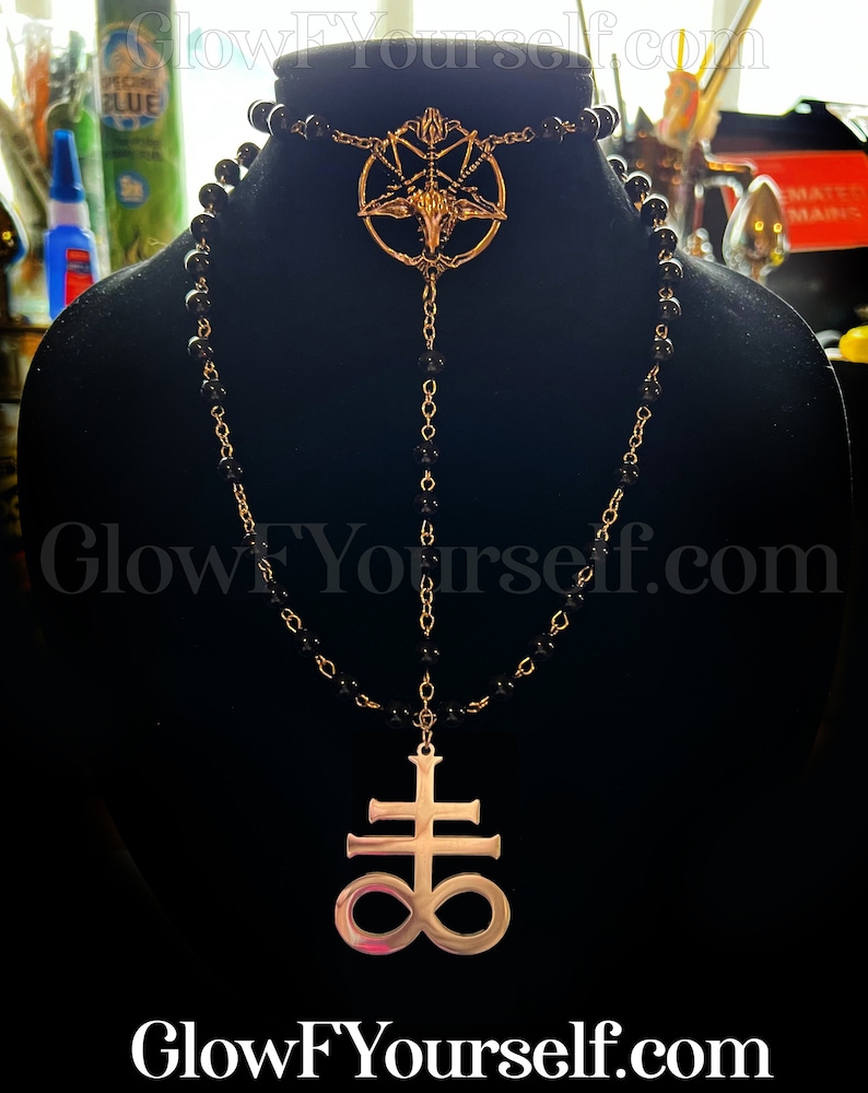 Op de afbeelding: Een zwarte kralenketting met twee gouden hangers. De bovenste hanger is een pentagram met een geitenkop in het midden. De onderste hanger is een kruis met een oneindigheidssymbool.