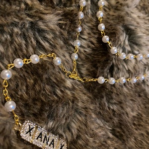 Pode incluir: Um colar de corrente dourada com pérolas de imitação e um pingente retangular. O pingente é cravejado com strass transparentes e apresenta a palavra "XANAX" em letras maiúsculas pretas. O colar é exibido em uma superfície marrom e peluda.