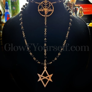 Op de afbeelding: Een zwarte kralenketting met twee gouden hangers. De bovenste hanger is een pentagram met een geitenkop in het midden. De onderste hanger is een zespuntige ster met een dubbele lijndesign.