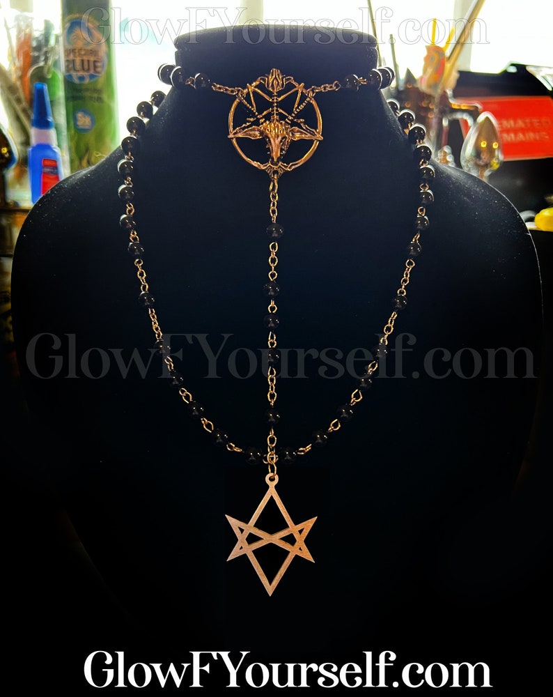 Op de afbeelding: Een zwarte kralenketting met twee gouden hangers. De bovenste hanger is een pentagram met een geitenkop in het midden. De onderste hanger is een zespuntige ster met een dubbele lijndesign.