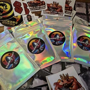 May include: Four iridescent holographic bags with a circular sticker on each. The stickers feature a cartoon illustration of a creature with the text "Thanks a ton!!" printed on each.