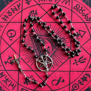 May include: A rosary-style necklace featuring black beads linked by a silver-toned metal chain. The centrepiece is a circular pendant depicting a goat head within a pentagram, and the end features a silver-toned crucifix with a figure.
