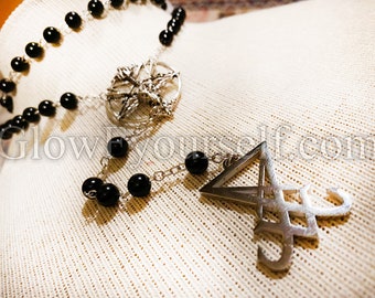Könnte beinhalten: Eine schwarze Perlenkette mit einem silbernen Anhänger, der ein Pentagramm und ein Symbol zeigt. Die Kette liegt auf einem weißen Stoffhintergrund.
