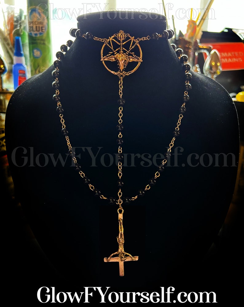 Op de afbeelding: Een zwarte kralenketting met twee gouden hangers. De bovenste hanger is een pentagram met een geitenkop in het midden. De onderste hanger is een gouden omgekeerd kruis met een figuur erop.