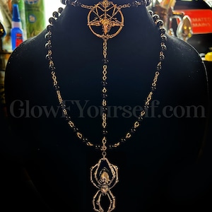 Op de afbeelding: Een zwarte kralenketting met een gouden hanger met een schedel en spin ontwerp. De ketting heeft ook een gouden hanger met een pentagram en een geitenkop.