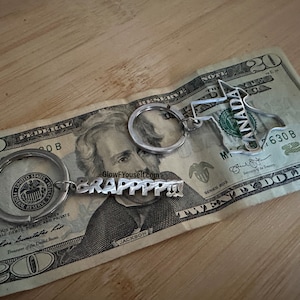 May include: A silver keychain with two charms. One charm is a cutout of the state of Canada and the other charm is a nameplate that reads "BRAPPPP". The keychain is resting on a twenty dollar bill.