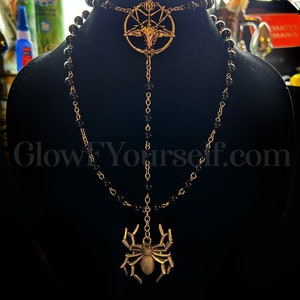 Op de afbeelding: Een zwarte kralenketting met een gouden hanger met een pentagram met een geitenkop in het midden. Een tweede gouden hanger in de vorm van een spin hangt aan de onderkant van de ketting.
