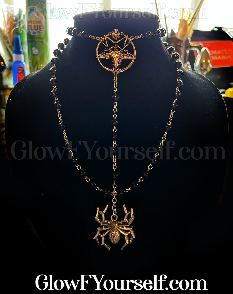 Op de afbeelding: Een zwarte kralenketting met een gouden hanger met een pentagram met een geitenkop in het midden. Een tweede gouden hanger in de vorm van een spin hangt aan de onderkant van de ketting.