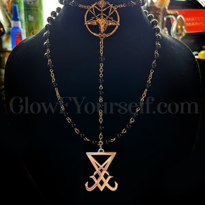 Op de afbeelding: Een zwarte kralenketting met twee gouden hangers. De bovenste hanger is een pentagram met een geitenkop in het midden. De onderste hanger is een zegel met een gestileerde 'X' en twee gebogen lijnen.