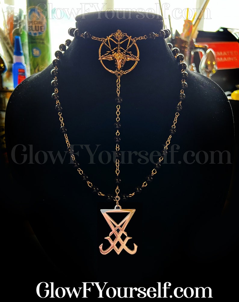 Op de afbeelding: Een zwarte kralenketting met twee gouden hangers. De bovenste hanger is een pentagram met een geitenkop in het midden. De onderste hanger is een zegel met een gestileerde 'X' en twee gebogen lijnen.