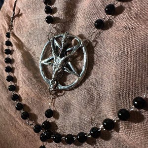 May include: A necklace featuring a silver-toned circular pendant with a goat head design centered within a pentagram. The pendant is connected to a chain composed of smooth, round, black beads linked together with thin silver-toned metal wire.