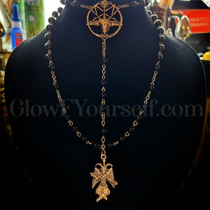 Op de afbeelding: Een zwarte kralenketting met twee gouden hangers. De bovenste hanger is een pentagram met een geitenkop in het midden. De onderste hanger is een gouden beeldje van een geitenachtige demon met vleugels. De ketting wordt gepresenteerd op een zwarte fluwelen standaard.