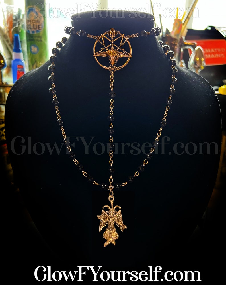 Op de afbeelding: Een zwarte kralenketting met twee gouden hangers. De bovenste hanger is een pentagram met een geitenkop in het midden. De onderste hanger is een gouden beeldje van een geitenachtige demon met vleugels. De ketting wordt gepresenteerd op een zwarte fluwelen standaard.