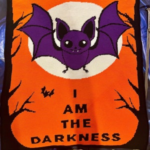 May include: A crocheted blanket with an orange background and a black border. A purple bat with black wings is flying in front of a white moon. The text "I AM THE DARKNESS" is written in black letters on the orange background.
