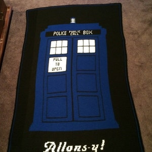 May include: A blue and black crocheted blanket featuring a TARDIS, a time-traveling police box from the Doctor Who television series. The blanket has the words "Police Public Call Box" and "Pull to Open" on the TARDIS, and the words "Allons-y!" at the bottom.