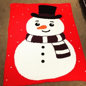 May include: A red crocheted blanket with a white snowman design. The snowman has a black top hat, a black and white striped scarf, and black eyes and buttons.