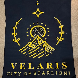 May include: A navy blue crocheted blanket with a yellow design featuring a mountain, a crescent moon, a star, and the text "VELARIS CITY OF STARLIGHT".