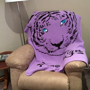 May include: A purple crocheted blanket with a black and white tiger face design. The tiger has blue eyes and the blanket says "Tiger" in black lettering.