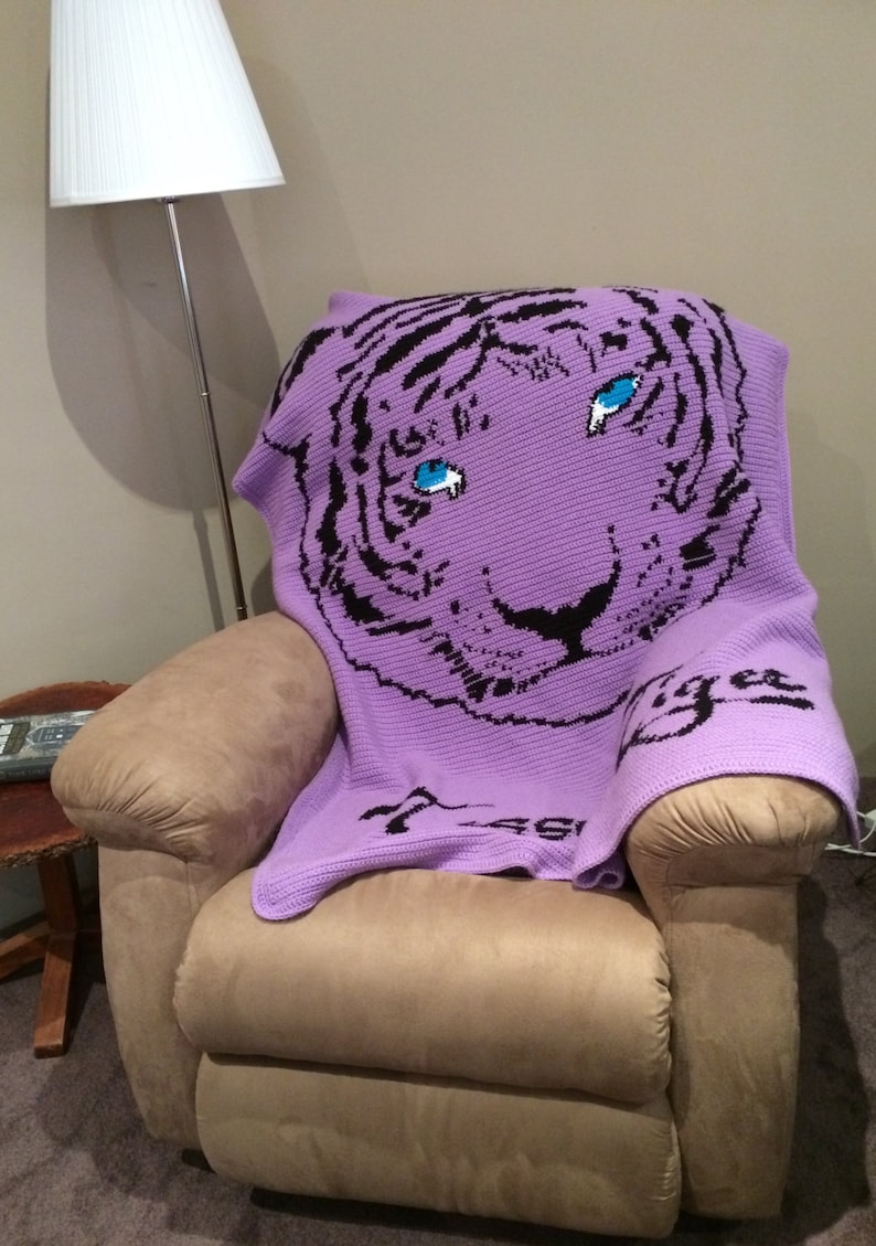 May include: A purple crocheted blanket with a black and white tiger face design. The tiger has blue eyes and the blanket says "Tiger" in black lettering.