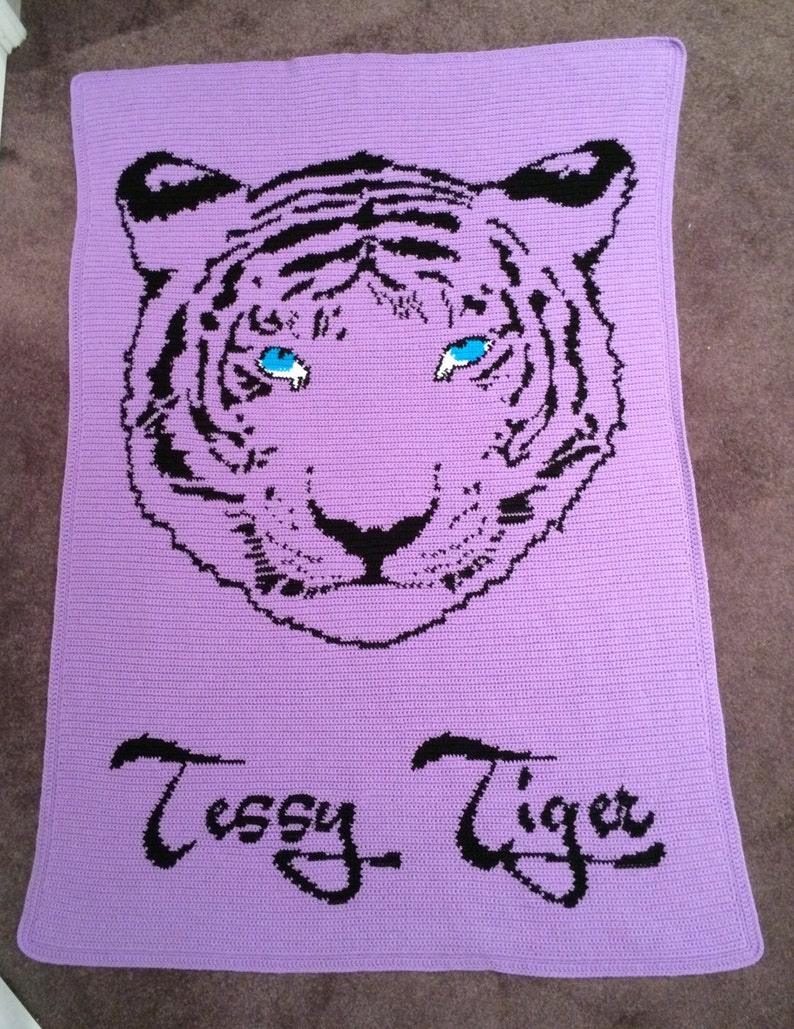 May include: A purple crocheted blanket with a black and white tiger face design. The tiger has blue eyes and the blanket is signed "Tessy Tiger".