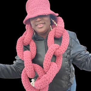 Puede incluir: Un conjunto de sombrero y bufanda de punto de color burdeos. El sombrero tiene un ala ancha y la bufanda es de estilo cadena gruesa.