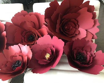 May include: A collection of six large, burgundy paper flowers. The flowers are made of multiple layers of paper and have a realistic appearance. The flowers are arranged on a white surface.
