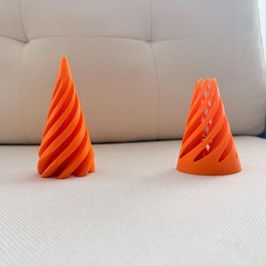 May include: Two bright orange, cone-shaped objects with a spiral, open-lattice design. The object on the left is taller and pointed at the top, while the object on the right is shorter with a flat, open top. Both feature a repeating twisted pattern.