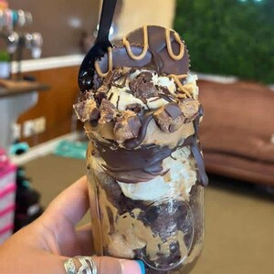 May include: A glass mason jar filled with layers of chocolate brownie, peanut butter, and vanilla ice cream. The dessert is topped with chocolate syrup, peanut butter drizzle, chopped chocolate candy pieces, and a large chocolate peanut butter cup.