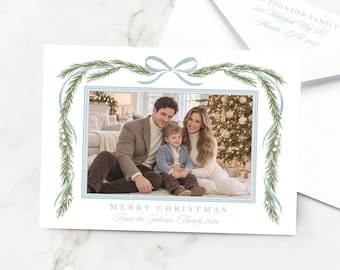 Peut inclure: Une carte de vœux de Noël blanche avec une photographie centrale d'une famille de trois personnes, encadrée par des branches de pin vertes et un ruban bleu clair. Le texte indique MERRY CHRISTMAS From the Johnson Family 2026, enveloppe incluse.