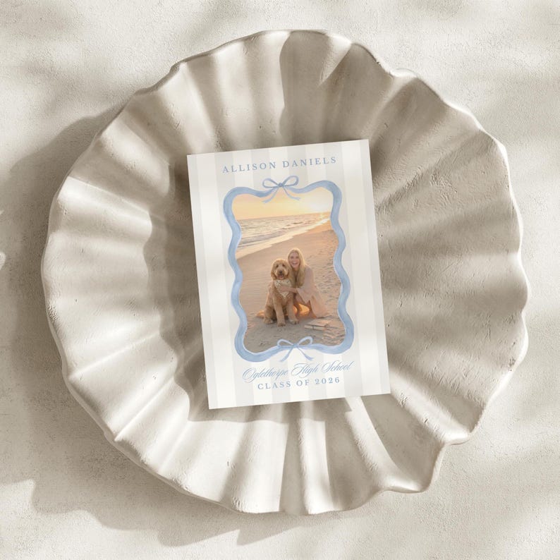 May include: A graduation announcement card featuring a photograph of a person and a dog on a beach, framed by a light blue border with bow accents. The card has vertical stripes and text that reads "Allison Daniels" and "Oglethorpe High School CLASS OF 2026."