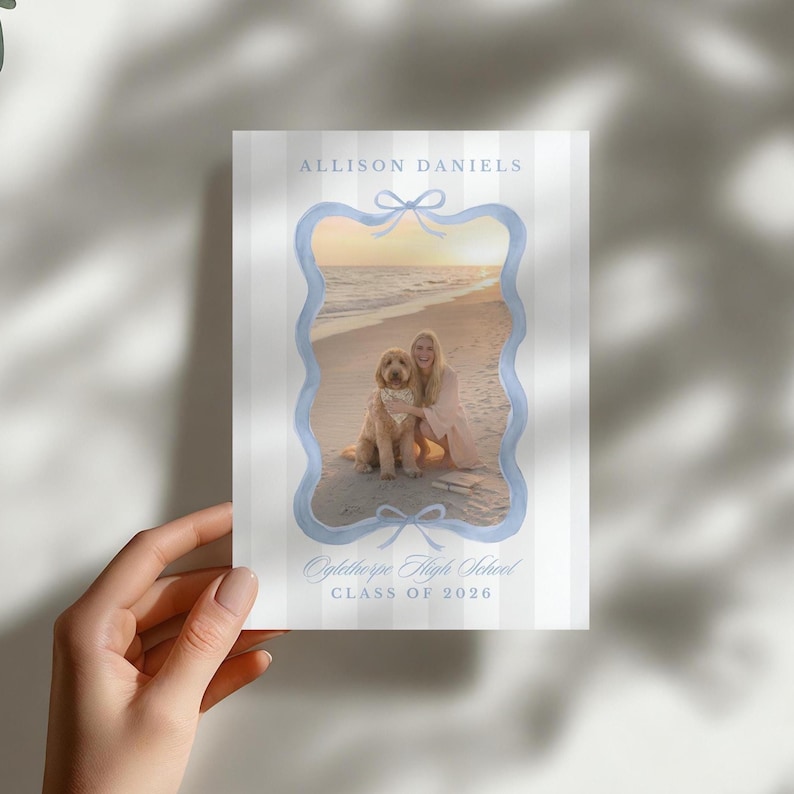 May include: A graduation announcement featuring a beach scene with a woman and a dog inside a light blue frame. The announcement has the name "Allison Daniels" at the top and "Oglethorpe High School, Class of 2026" at the bottom.