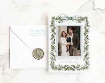 Può includere: Un biglietto di Natale bianco con una fotografia centrale di tre persone, incorniciata da un'illustrazione ad acquerello di rami di pino verdi e un nastro azzurro. Il testo recita 2026, MERRY CHRISTMAS e From the Miller Family. Include una busta abbinata con sigillo in ceralacca.