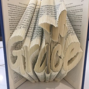 May include: A book art sculpture of the word "Grace" created by folding the pages of a book. The sculpture is made from a book with a blue cover and the pages are folded to create the letters of the word.