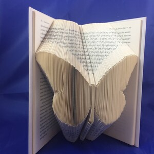 May include: A book folded into the shape of a butterfly. The book is open and the pages are folded to create the wings of the butterfly. The butterfly is made of paper and is a light brown color.
