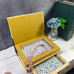 May include: A book-shaped box with a hidden compartment, lined with floral fabric. The box is open, revealing a pink pearl necklace inside. The box is sitting on a white wooden surface.