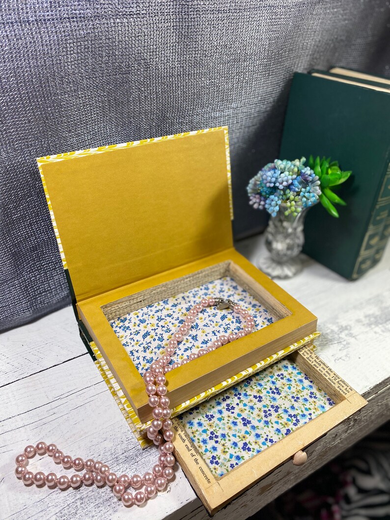 May include: A book-shaped box with a hidden compartment, lined with floral fabric. The box is open, revealing a pink pearl necklace inside. The box is sitting on a white wooden surface.