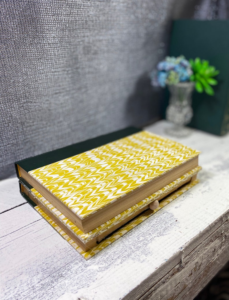 May include: A yellow and white patterned book with a hidden drawer. The book is closed and sitting on a white wooden surface.