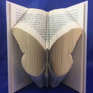 May include: A book art sculpture of a butterfly made by folding the pages of a book. The butterfly is made of cream-colored paper and is standing on a blue surface.