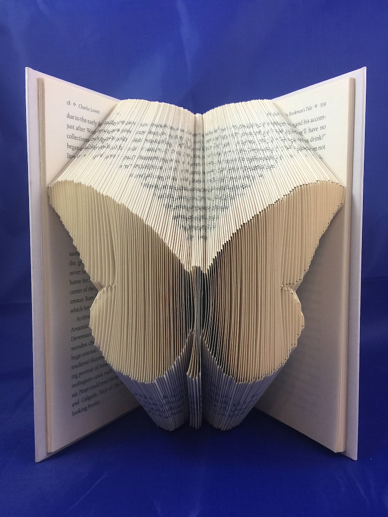 May include: A book art sculpture of a butterfly made by folding the pages of a book. The butterfly is made of cream-colored paper and is standing on a blue surface.