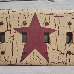 May include: A double light switch cover with a distressed, beige background and a red star design. The cover has six screws.