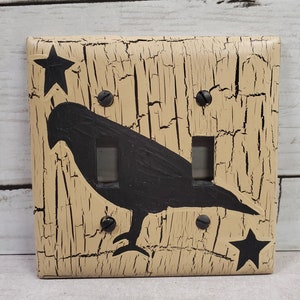 May include: A light beige double light switch cover with a black silhouette of a crow and two black stars. The cover has a distressed, crackled paint finish.