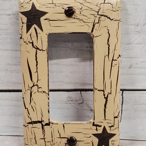 May include: A light beige light switch cover with a distressed, cracked paint finish. Two black stars are painted on the cover, one on each side of the switch opening. Two dark brown screws are visible on the cover.