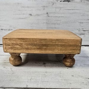 May include: A small, square, wooden riser with a natural finish. The riser is supported by four rounded, ball-shaped feet. The wood grain is visible, adding texture to the piece. This decorative item is suitable for displaying small objects.